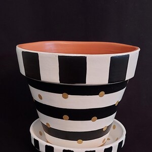 May include: A decorative flower pot with a matching saucer. The pot is painted with alternating black and white stripes and gold polka dots. The rim of the pot is painted black and white stripes. The interior of the pot is a terracotta color.