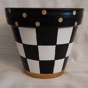 May include: A hand-painted ceramic flower pot. The top rim is black with gold polka dots. The body features a black and white checkerboard pattern. The base is a light tan color. The pot is designed for indoor or outdoor use.