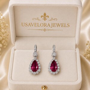 Può includere: Un paio di orecchini pendenti color argento con gemme rosse a forma di pera circondate da piccole pietre trasparenti, presentati in un portagioie in velluto color crema con il testo USAVELORA JEWELS stampato in oro sul coperchio interno.