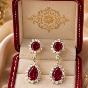 Puede incluir: Un par de pendientes colgantes de oro con una piedra redonda de color rubí en la parte superior y una piedra en forma de pera de color rubí en la parte inferior, rodeadas por un halo de pequeñas piedras claras, en un joyero de terciopelo rojo.