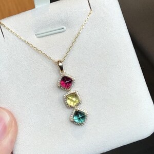 Könnte beinhalten: Eine goldene Halskette mit einem vertikalen Anhänger, der drei quadratische Edelsteine in Magenta, Gelb und Türkis enthält, die jeweils von einem Kranz aus kleinen weißen Steinen umgeben sind. Der Anhänger hängt an einer weißen Präsentationskarte.