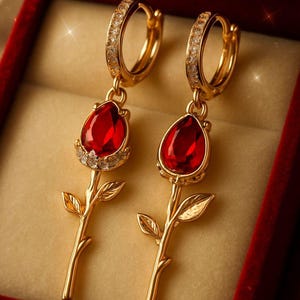 May include: A pair of gold-colored earrings shaped like roses. Each earring features a red, teardrop-shaped gemstone at the flower's center, surrounded by small, clear stones. The earrings are displayed in a red velvet jewelry box.