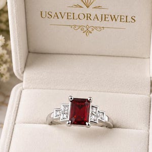 May include: A silver-toned ring featuring a rectangular red centre stone flanked by two smaller square white stones on each side. The ring is displayed inside a cream-coloured velvet box with the gold text USAVELORAJEWELS printed on the inner lid.