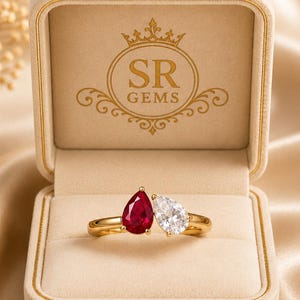 May include: A gold ring featuring two pear-cut stones, one deep red ruby and one clear white diamond, set in a bypass style. The ring is displayed inside a beige velvet jewellery box with the SR GEMS logo printed in gold on the inner lid.