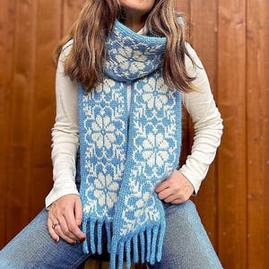 May include: A blue and white knitted scarf with a floral pattern and fringe. The scarf is worn by a person with long brown hair.