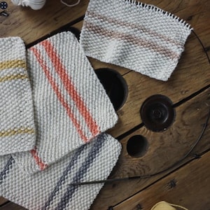 May include: Four knitted squares in white with different coloured stripes. The squares are made with a textured stitch and have stripes of orange, yellow, grey, and pink.
