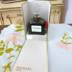 May include: A vintage perfume bottle of Réplique by Raphael Paris, presented in a gold-trimmed box. The dark glass bottle has a tulip-shaped stopper and a label with the brand name. The box is open, revealing the bottle on a white platform.