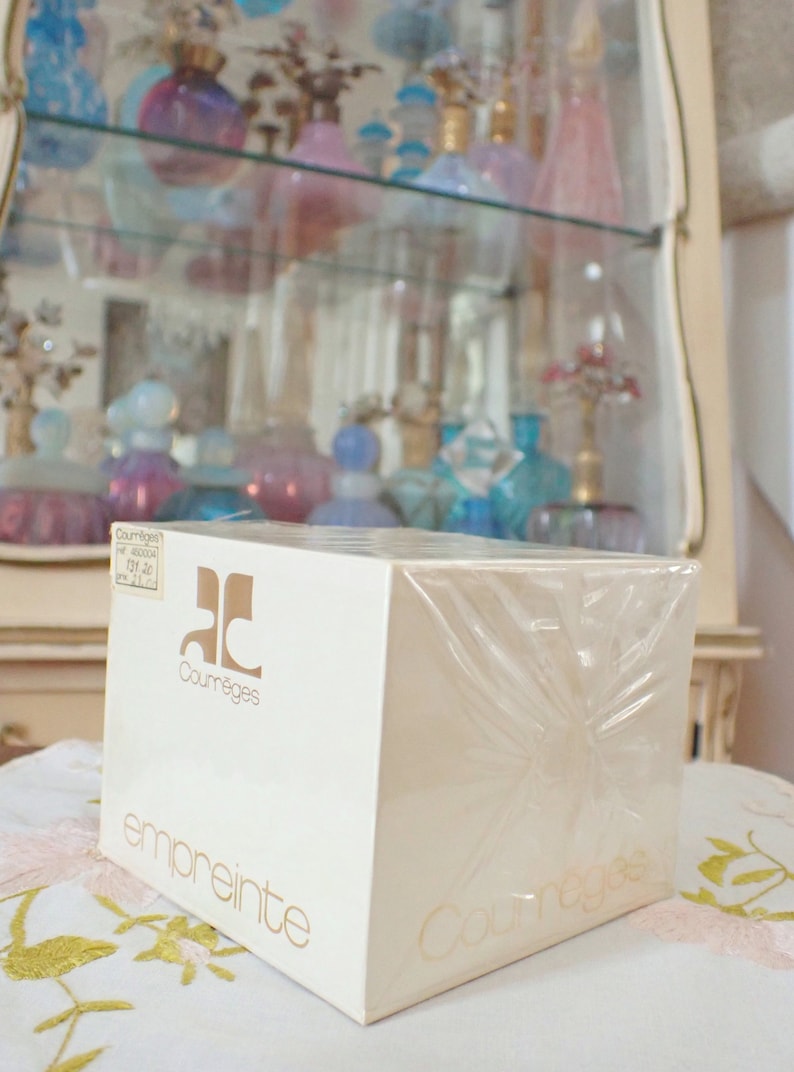 May include: A white box with a clear plastic wrap, featuring the text "Courr&egrave;ges" and "empreinte" in gold lettering. The box is sitting on a white surface with a floral pattern.