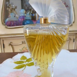 Puede incluir: Un frasco de perfume de vidrio transparente con un tap&oacute;n en forma de abanico. La botella tiene un dise&ntilde;o &uacute;nico y retorcido y est&aacute; llena de l&iacute;quido dorado. El tap&oacute;n es esmerilado y el cuello de la botella es dorado. Sobre un mantel blanco con bordado floral rosa.