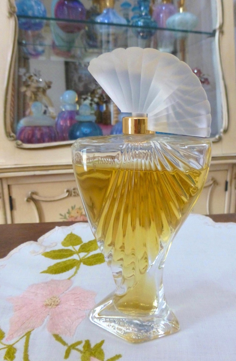 Puede incluir: Un frasco de perfume de vidrio transparente con un tap&oacute;n en forma de abanico. La botella tiene un dise&ntilde;o &uacute;nico y retorcido y est&aacute; llena de l&iacute;quido dorado. El tap&oacute;n es esmerilado y el cuello de la botella es dorado. Sobre un mantel blanco con bordado floral rosa.