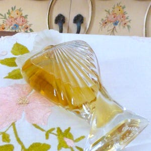 Puede incluir: Un frasco de perfume vintage de vidrio transparente, con un dise&ntilde;o en forma de abanico. La botella contiene un l&iacute;quido dorado y tiene una base clara y angular. Descansa sobre una tela blanca con bordados florales rosas.