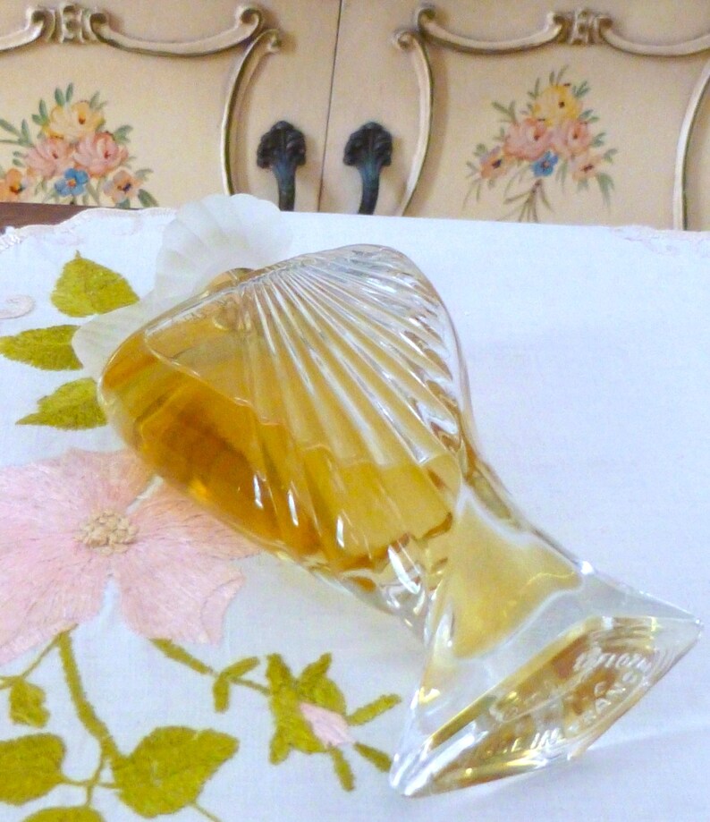 Puede incluir: Un frasco de perfume vintage de vidrio transparente, con un dise&ntilde;o en forma de abanico. La botella contiene un l&iacute;quido dorado y tiene una base clara y angular. Descansa sobre una tela blanca con bordados florales rosas.