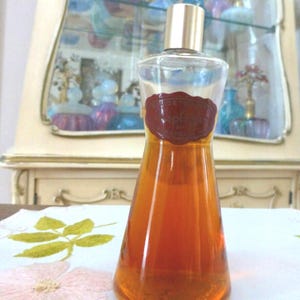 May include: A clear glass perfume bottle with a gold cap and a dark red label. The bottle is filled with a golden-brown liquid. The bottle is on a white tablecloth with floral embroidery.