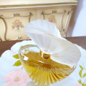 Puede incluir: Un frasco de perfume de vidrio transparente con un l&iacute;quido dorado en su interior. El frasco tiene una forma &uacute;nica y est&aacute; coronado con un tap&oacute;n blanco en forma de abanico. Se encuentra sobre una tela blanca con bordados florales rosas. El fondo muestra un mueble color crema.