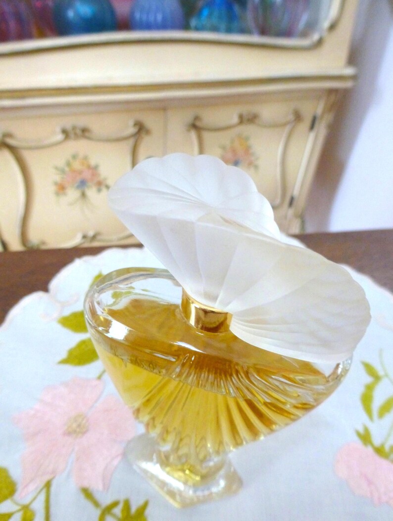 Puede incluir: Un frasco de perfume de vidrio transparente con un l&iacute;quido dorado en su interior. El frasco tiene una forma &uacute;nica y est&aacute; coronado con un tap&oacute;n blanco en forma de abanico. Se encuentra sobre una tela blanca con bordados florales rosas. El fondo muestra un mueble color crema.