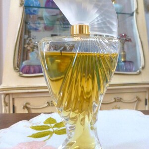 Puede incluir: Un frasco de perfume vintage con un dise&ntilde;o de vidrio transparente esculpido. El frasco tiene un tap&oacute;n en forma de abanico y un cuello dorado. El l&iacute;quido del interior es de color dorado. El frasco est&aacute; sobre un mantel blanco con bordados florales.
