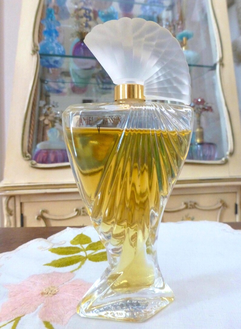 Puede incluir: Un frasco de perfume vintage con un dise&ntilde;o de vidrio transparente esculpido. El frasco tiene un tap&oacute;n en forma de abanico y un cuello dorado. El l&iacute;quido del interior es de color dorado. El frasco est&aacute; sobre un mantel blanco con bordados florales.