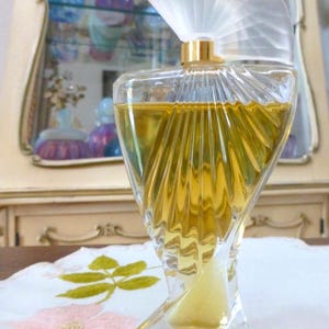 Puede incluir: Un frasco de perfume de vidrio transparente con un tap&oacute;n en forma de abanico y detalles dorados. El frasco tiene un dise&ntilde;o retorcido &uacute;nico y est&aacute; lleno de un l&iacute;quido dorado. El fondo incluye un armario con otros frascos de perfume.
