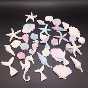 Könnte beinhalten: Eine Sammlung von weißen, rosa und blauen Fondant-Dekorationen zum Thema Meer. Die Dekorationen umfassen Muscheln, Seesterne, Seepferdchen und Meerjungfrauen-Schwänze.