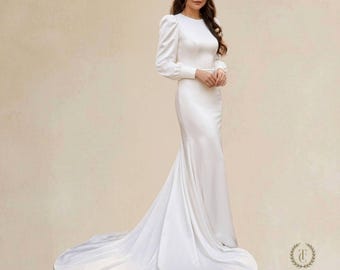 May include: A floor-length white gown with long sleeves, a round neckline, and puff shoulders. The dress features a fitted bodice that transitions into a flared skirt with a train. The brand name Tehila Chaya is printed in the bottom right corner.