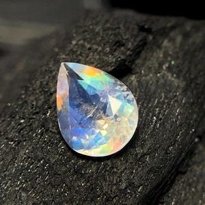 May include: A pear-shaped iridescent gemstone with a rainbow sheen. The gemstone is set against a dark background.