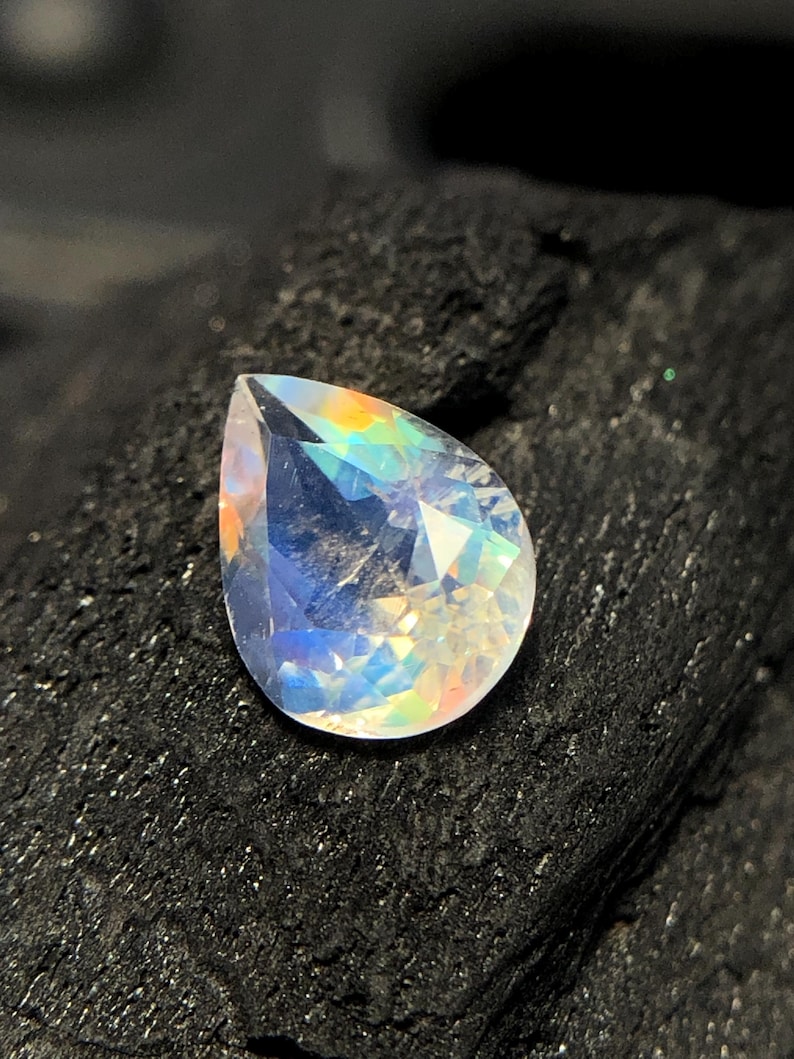 May include: A pear-shaped iridescent gemstone with a rainbow sheen. The gemstone is set against a dark background.