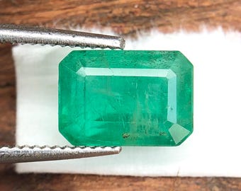 May include: A vibrant, rectangular emerald gemstone, held by silver tweezers. The gem is a deep, rich green with visible facets, set against a blurred wooden background. The emerald's clarity and cut are highlighted in this close-up shot.