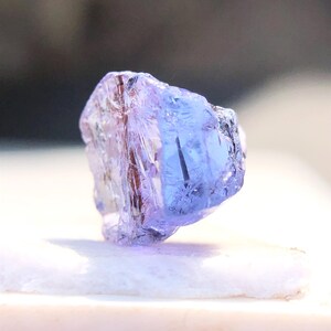 May include: A translucent purple gemstone with a faceted cut. The gemstone is on a white surface.