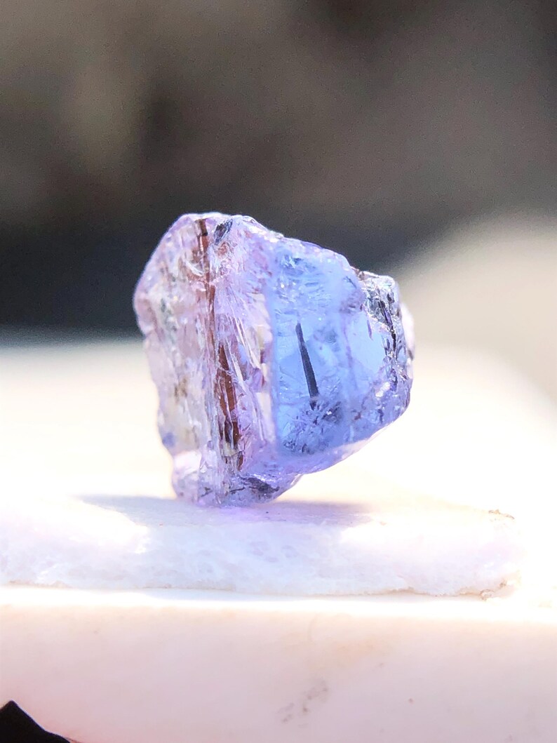 May include: A translucent purple gemstone with a faceted cut. The gemstone is on a white surface.