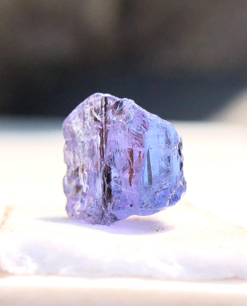 May include: A faceted, purple-blue gemstone with a clear, glassy appearance. The gemstone is rough-cut and has a natural, irregular shape.