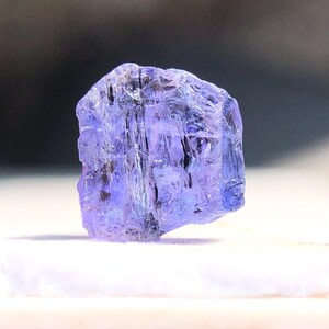 May include: A faceted, purple-blue gemstone with a rough, natural finish. The gemstone is translucent and has a slightly iridescent sheen.
