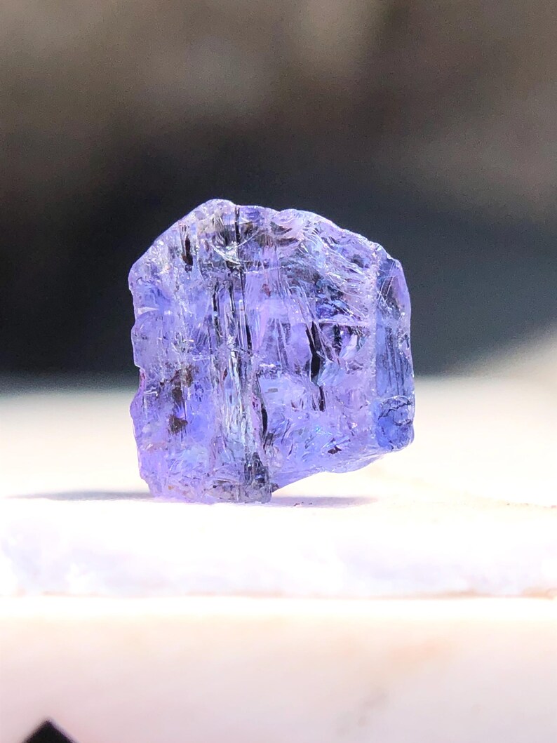 May include: A faceted, purple-blue gemstone with a rough, natural finish. The gemstone is translucent and has a slightly iridescent sheen.