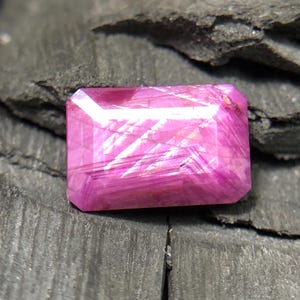 May include: A rectangular, faceted pink gemstone with visible internal inclusions and a polished surface. The stone displays a vibrant magenta hue with light reflecting off its multiple angled facets, showcasing a clear, crystalline structure.