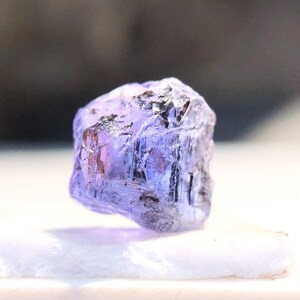 May include: A faceted, purple gemstone with a rough, natural finish. The gemstone is translucent and has a slight sheen.
