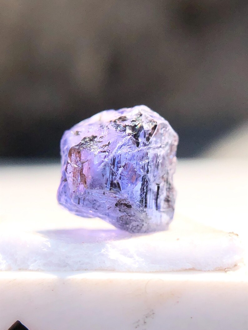 May include: A faceted, purple gemstone with a rough, natural finish. The gemstone is translucent and has a slight sheen.