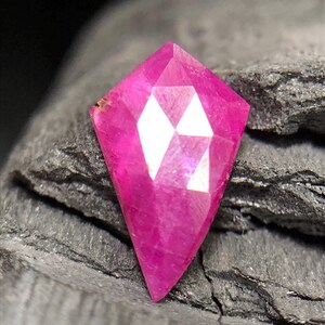 Puede incluir: Una piedra preciosa facetada en forma de cometa con un color rosa magenta vibrante y saturado. La piedra presenta un corte geométrico con múltiples facetas triangulares y en forma de diamante que reflejan la luz, creando una silueta afilada y puntiaguda en la base.