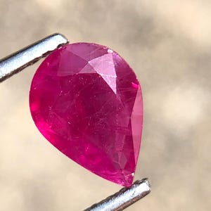 Pode incluir: Uma gema de rubi facetada em forma de pêra, segurada por pinças de prata. O rubi é de uma cor vermelha profunda e vibrante, com inclusões internas visíveis. A gema é cortada para maximizar a reflexão da luz.