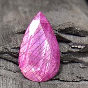 Puede incluir: Una piedra preciosa facetada en forma de pera de un vibrante color rosa magenta. La piedra presenta un patrón interno distintivo de líneas finas entrecruzadas y reflejos de luz en su superficie, mostrando una calidad translúcida.