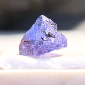 May include: A purple gemstone with a rough, faceted surface. The gemstone is triangular in shape and has a light purple hue.