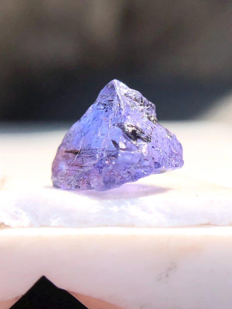 May include: A purple gemstone with a rough, faceted surface. The gemstone is triangular in shape and has a light purple hue.