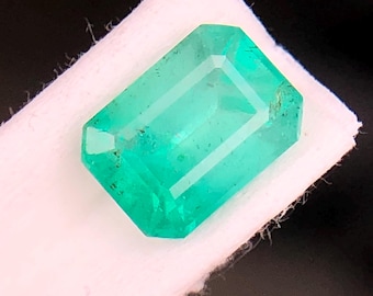 May include: A rectangular emerald gemstone with a vibrant green hue. The gem is faceted, showcasing internal lines and a polished surface. It rests on a white, textured fabric against a dark background.