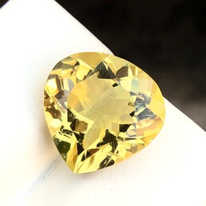 May include: A heart-shaped yellow gemstone with a faceted cut. The gemstone is on a white surface.