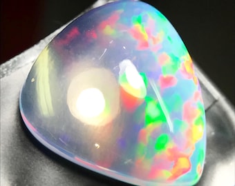 May include: A triangular, iridescent opal gemstone. The opal displays a play of color with flashes of red, green, yellow, and blue. The stone is translucent with a milky white base, and is set against a dark background.