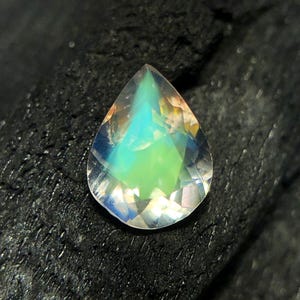 May include: A teardrop-shaped iridescent gemstone with a pearlescent finish. The gemstone is a light blue color with hints of green and yellow.