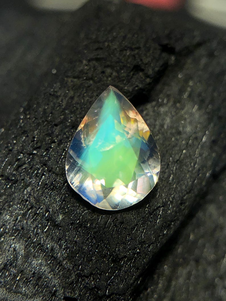 May include: A teardrop-shaped iridescent gemstone with a pearlescent finish. The gemstone is a light blue color with hints of green and yellow.