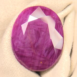 May include: An oval-shaped, faceted gemstone with a deep magenta and purple hue. The stone features a distinct internal fibrous texture and a polished surface with multiple triangular facets that reflect light across its translucent body.