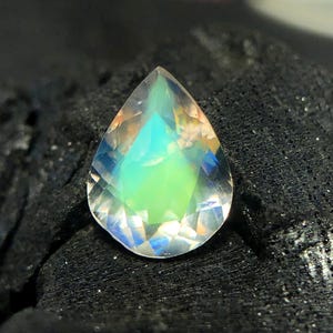 May include: A teardrop-shaped iridescent gemstone with a green and blue sheen. The gemstone is faceted and has a clear, glassy appearance.