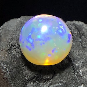 May include: A round, iridescent opal gemstone with a polished surface. The opal displays a mix of colors, including blue, yellow, and white, with a play-of-color effect. The gemstone is set against a dark, textured background.