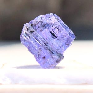 May include: A purple and blue gemstone with a faceted cut. The gemstone is a rough, natural crystal with a translucent appearance.