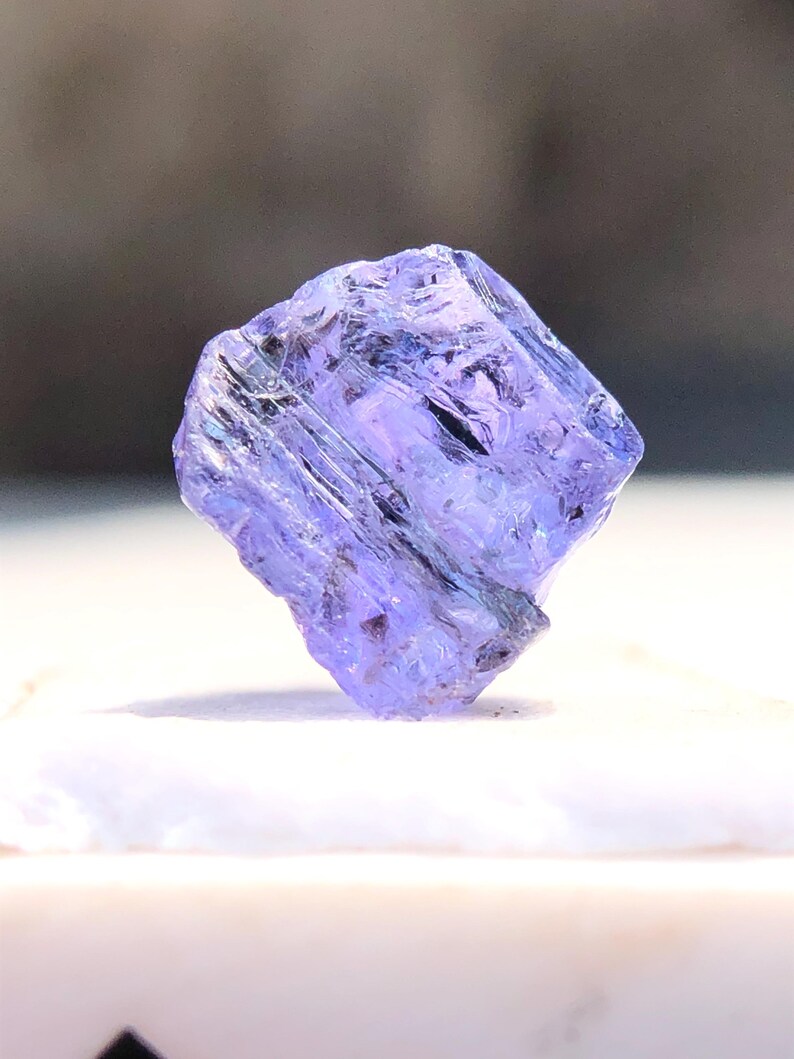 May include: A purple and blue gemstone with a faceted cut. The gemstone is a rough, natural crystal with a translucent appearance.